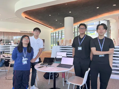Four people facing the camera with smiles standing around a white table with three laptops on it. Two people are standing to the left of the table and two people are standing to the right of the table.