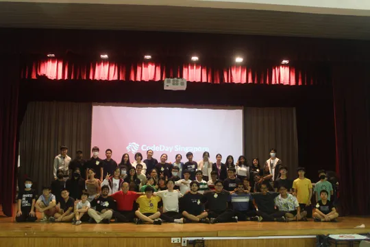 A wide shot of 50 people sitting on a stage with a screen behind them with the words "CodeDay Singapore" on it. The people are spread across three rows with the front row sitting on the stage floor.